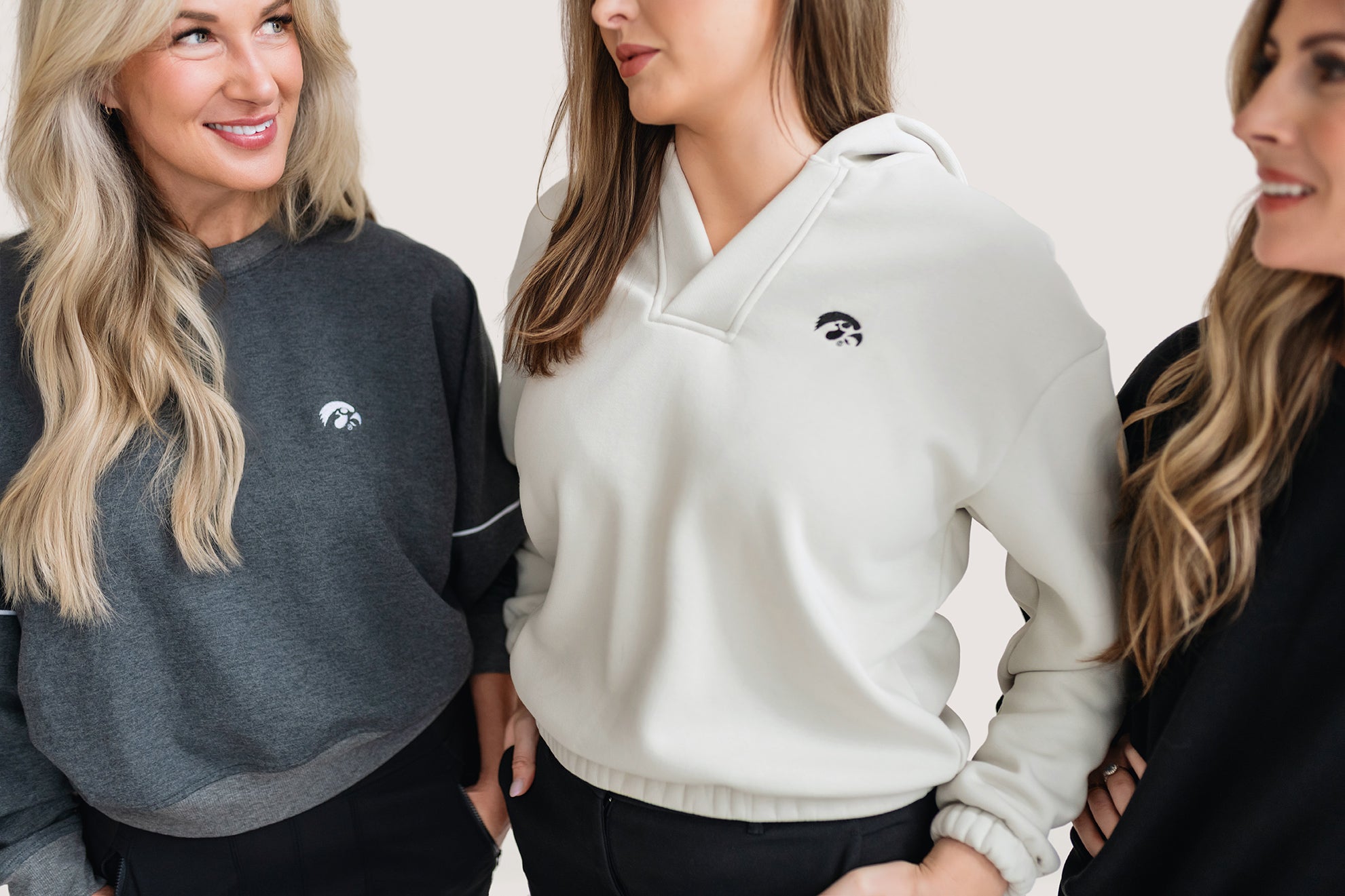 Three women wearing sweatshirts with logos, standing close together against a plain background.